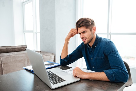 Picture of confused young man sitting near laptop and holding head with his hand. Looking at laptop.