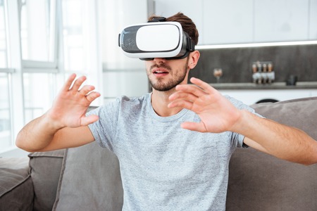 Image of young man dressed in grey t-shirt wearing virtual reality device while sitting on sofa