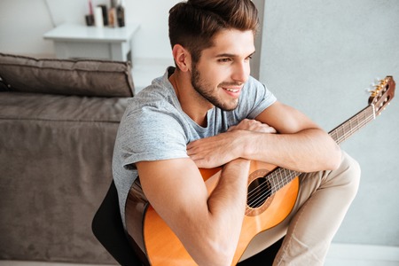 Picture of young man sitting with guitar on chair and look aside.