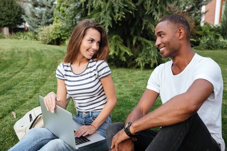 Happy multiethnic young couple sitting and using laptop on lawn outdoors