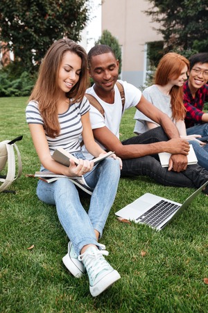 Multiethnic group of happy young people reading book and using laptop on lawn outdoors