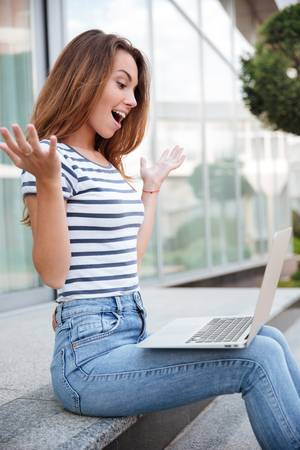 Happy excited young woman sitting and using laptop outdoorsの写真素材