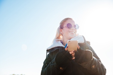 Smiling young girl in jacket holding mobile phone and looking away outdoorsの写真素材