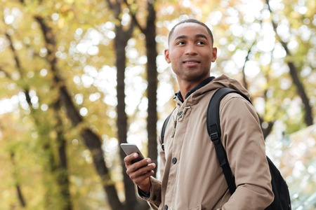 Photo of cheerful young african man holding his phone in hands and looking aside.