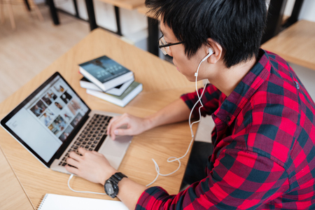Photo of asian male dressed in shirt in a cage and wearing glasses using laptop at the library and listen to music. Looking at laptop.の写真素材