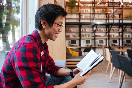 Cheerful asian man dressed in shirt in a cage and wearing glasses sitting at the library. Looking at...