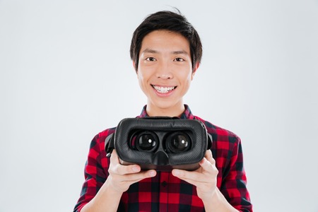 Photo of young asian man dressed in casual shirt in a cage and showing to camera virtual reality device. Isolated over white background.の写真素材