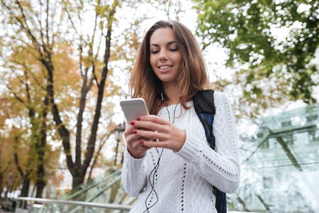 Happy cute young woman with earphones listening to music from mobile phone in park