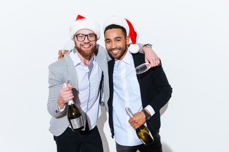 Two smiling young men in santa claus hats with bottle of champagne and glasses over white backgroundの写真素材