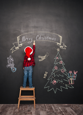 Picture of happy child wearing hat standing on stool near Christmas tree while drawing on blackboard.の写真素材