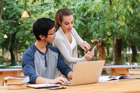 Smiling young student using laptop and mobile phone in outdoor cafe