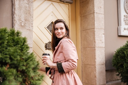 Pretty model in coat with coffee. stands sideways. on the street