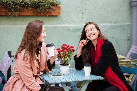Two smiling beautiful young women drinking coffee and talking in outdoor cafeの写真素材