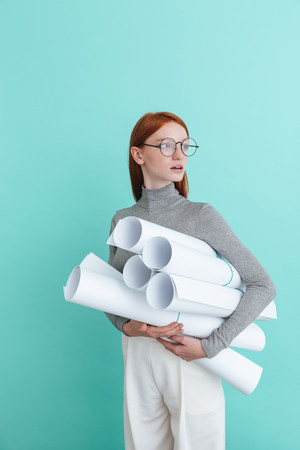 Pensive young woman in round glasses standing and holding blueprints over blue backgroundの写真素材