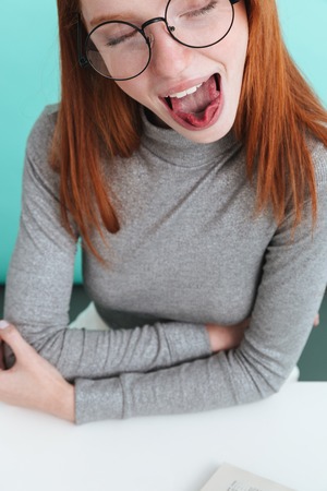 Closeup of funny young woman in round glasses sitting and showing tongue over blue backgroundの写真素材