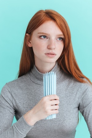 Portrait of serious redhead young woman holding pencils over blue backgroundの写真素材