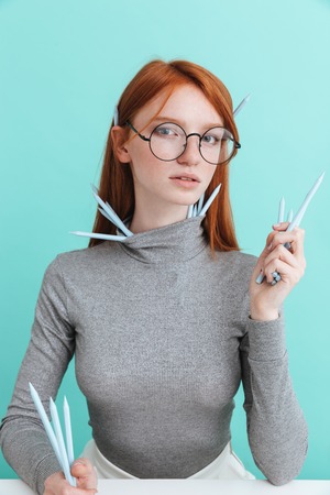 Portrait of beautiful young woman in roud glasses sitting and holding pencils over blue background