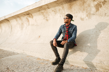 Photo of attractive african man wearing cap and watch while holding eyeglasses walking on the beach. Looking aside.の写真素材