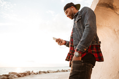 Young man walking on beach and chatting by his phone while listening music with earphones. Looking...