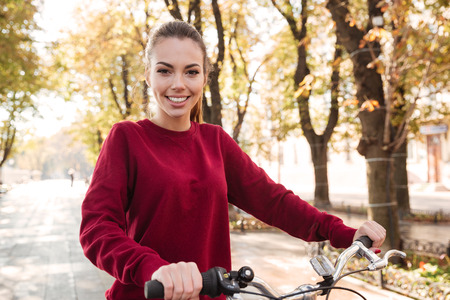 Image of cheerful lady dressed in sweater walking with her bicycle outdoors. Look at camera.