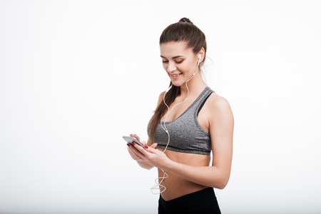 Portrait of a smiling fitness woman texting on mobile phone and wearing earphones isolated on a white backgroundの写真素材