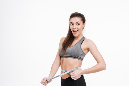 Portrait of a happy surprised sports woman holding tape measure around her waist isolated on a white...