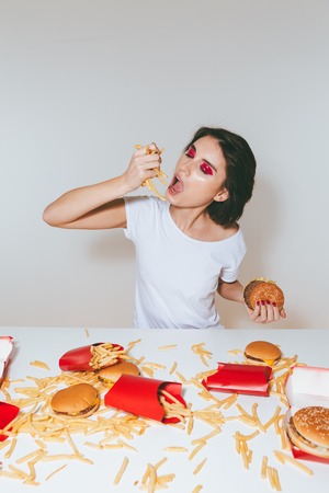 Pretty young woman sitting and eating french fries at the table with fast food over white background