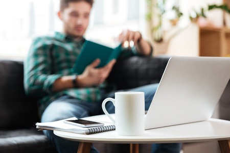 Man in green shirt reading book on sofa on blur background near the table with laptop in office. Focus on cup. Coworkingの写真素材