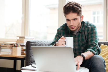 Man in green shirt with cup of tea in hand looking at laptop and sitting on sofa. Coworkingの写真素材