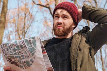 Portrait of confused bearded man wearing hat holding map in hands standing in the forest. Look aside.の写真素材