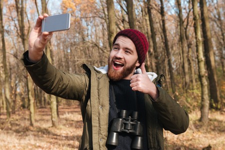 Image of cheerful bearded man wearing hat making selfie by the phone in the forest. Looking at phone and smile while making thumbs up gesture.の写真素材