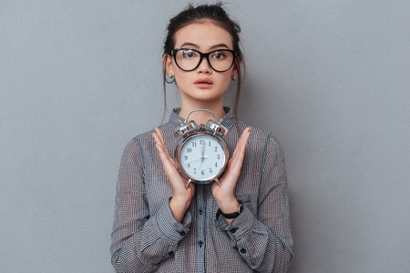 Young surprised asian woman in glasses and shirt holding the clock in hands. Isolated gray...