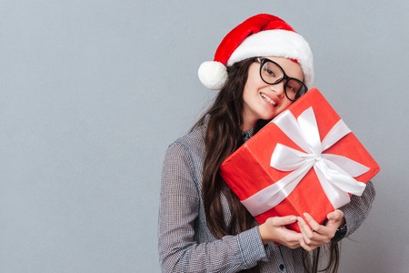 Smiling Asian girl in christmas hat holding gift in hands and looking at camera. Isolated gray...