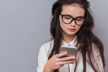 Serious pretty Asian woman in glasses writing message on phone. Front view. Isolated gray backgroundの写真素材
