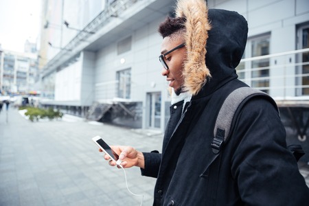 Image of african man holding his cellphone in hands and chatting while listen to music. Look at...