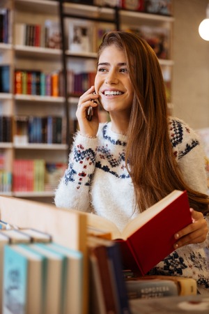 Smiling young woman holding book while talking on the phone in libraryの写真素材