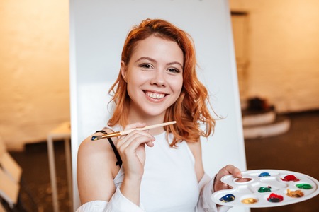 Picture of happy redhead young woman painter standing over blank canvas in artist workshop. Look at...