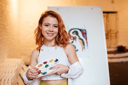 Image of happy young woman painter with red hair standing over blank canvas in artist workshop. Look at camera while holding palette and paintbrush.の写真素材