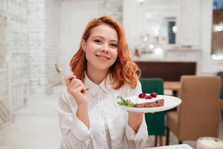 Image of redhead cheerful young woman eating cake in cafe. Looking at the camera.の写真素材