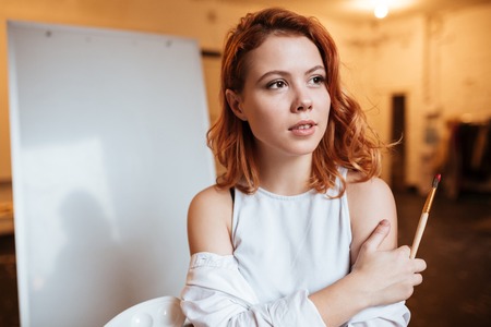 Photo of concentrated young lady painter with red hair standing over blank canvas in artist workshop. Look aside while holding palette and paintbrush.の写真素材