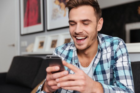 Image of young laughing man dressed in shirt in a cage print sitting on sofa in home and looking at...