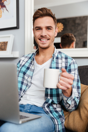 Picture of cheerful bristle man dressed in shirt in a cage print sitting on sofa in home and using laptop computer. Looking at camera while drinking tea.の写真素材