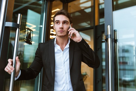 Serious business man in suit talking on phone in office and holding the door. From below imageの写真素材