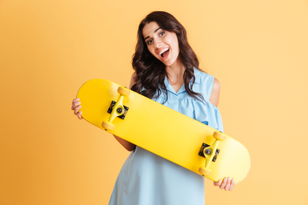 Portrait of a smiling excited brunette woman holing skateboard isolated on the orange backgroundの写真素材