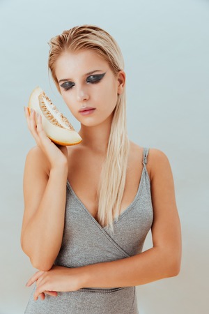 Attractive young woman with stylish makeup holding slice of melon over grey backgroundの写真素材