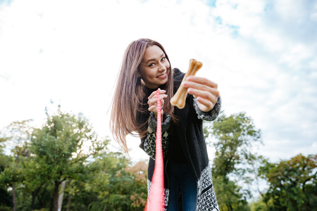 Smiling charming young woman showing and giving bone to her dog in parkの写真素材