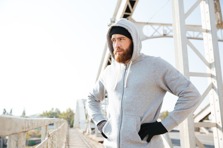 Portrait of a young sport athlete man in hoodie standing at the urban bridge with hands on hipsの写真素材