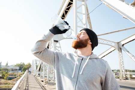 Portrait of a young bearded athlete in sportswear drinking water while standing at the bridgeの写真素材