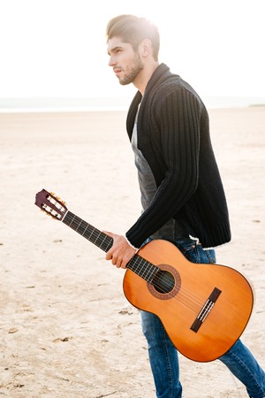 Portrait of a young casual man walking across seashore with guitar