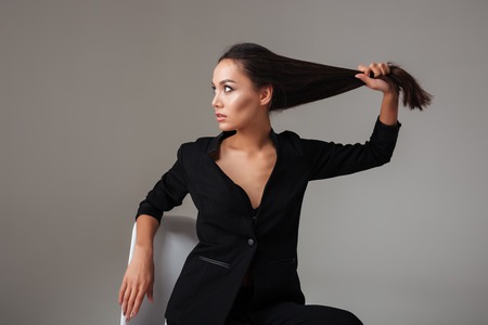 Portrait of a young brunette woman in black suit sitting and holding her long hair over gray backgroundの写真素材